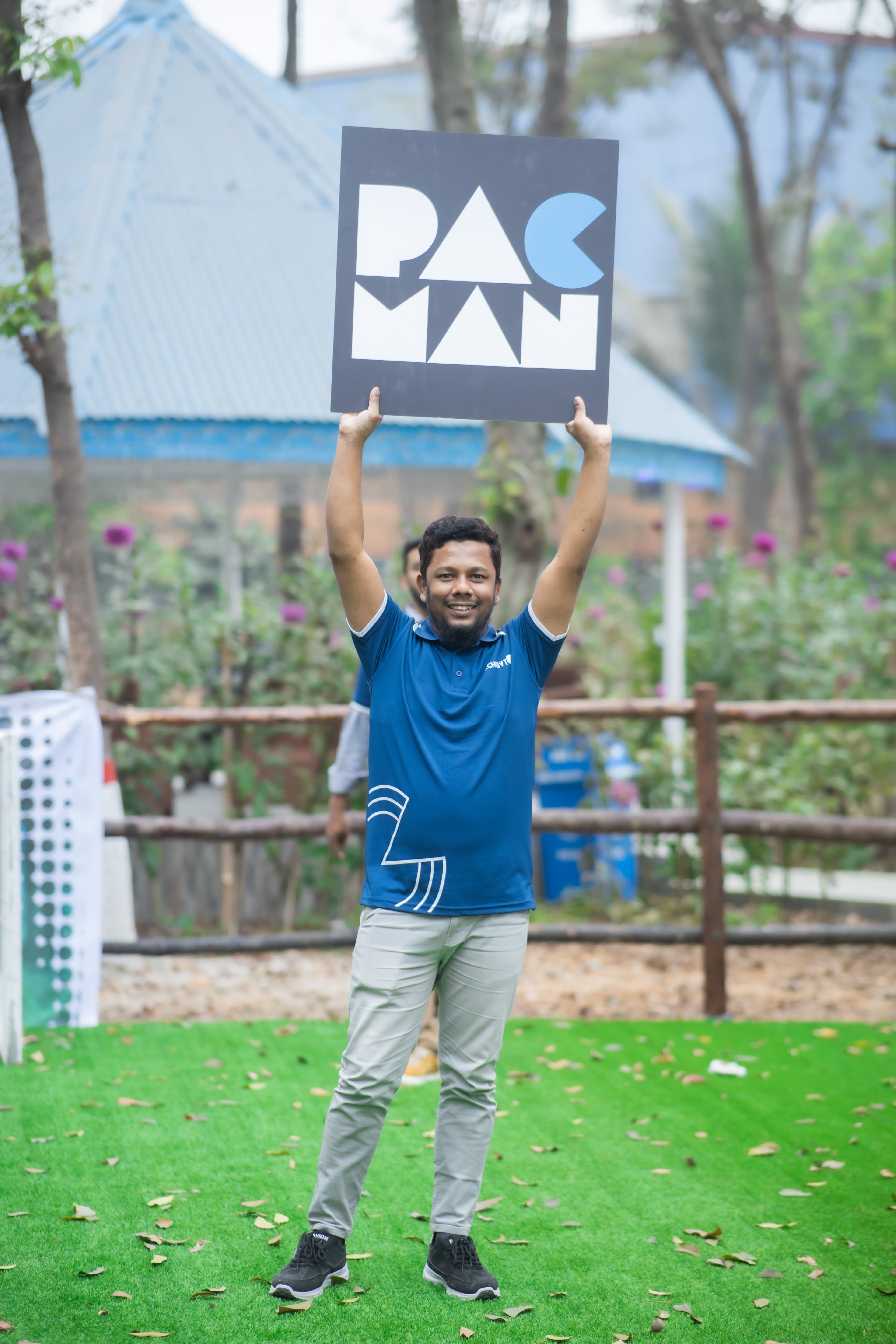 Rashed Ahmed Shaon smiling and holding a square “PAC MAN” sign above his head, standing on a lawn in a garden